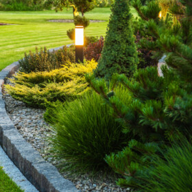 Landscaped Garden at the Backyard with Evenly Mowed Lawn and Island Bed with a Variety of Shrubs, Bushes, Trees and Evergreen Plants. Decorated with Bollard Outdoor Lighting Lamp.